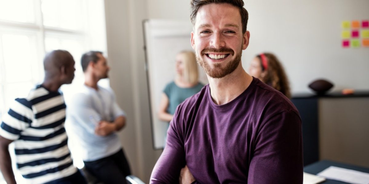 Smiling young designer standing confidently with his arms crossed after a boardroom meeting with colleagues standing in the background